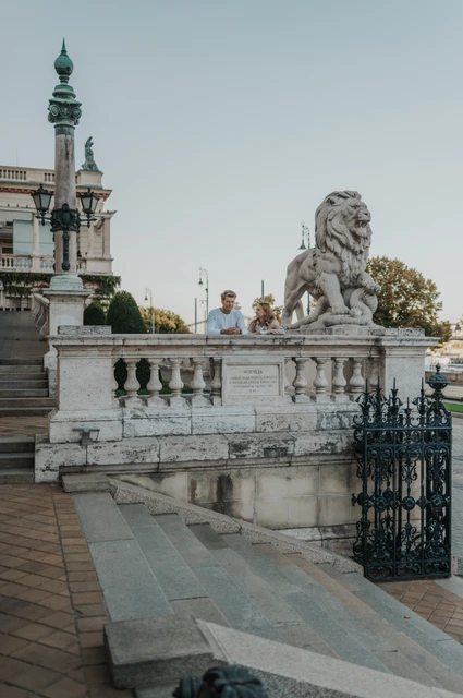 A city terrace beside the stone lion at a Budapest couple photoshoot A wide portrait of the couple behind a stone balustrade beside a large lion statue and ornate green lamp at Castle Garden Bazaar during their couple photoshoot in Budapest.