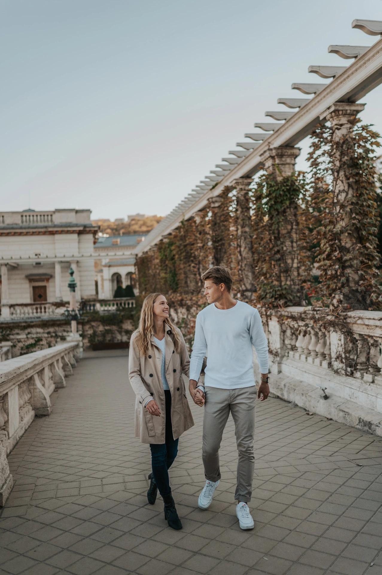 The couple walking towards the camera beside the ivy-covered pergola and stone railing at Castle Garden Bazaar during their couple photoshoot in Budapest.