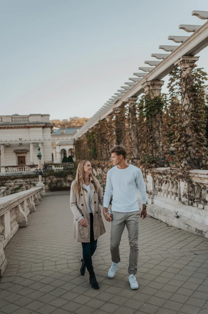 A relaxed walk along the garden terrace at a Budapest couple photoshoot The couple walking towards the camera beside the ivy-covered pergola and stone railing at Castle Garden Bazaar during their couple photoshoot in Budapest.