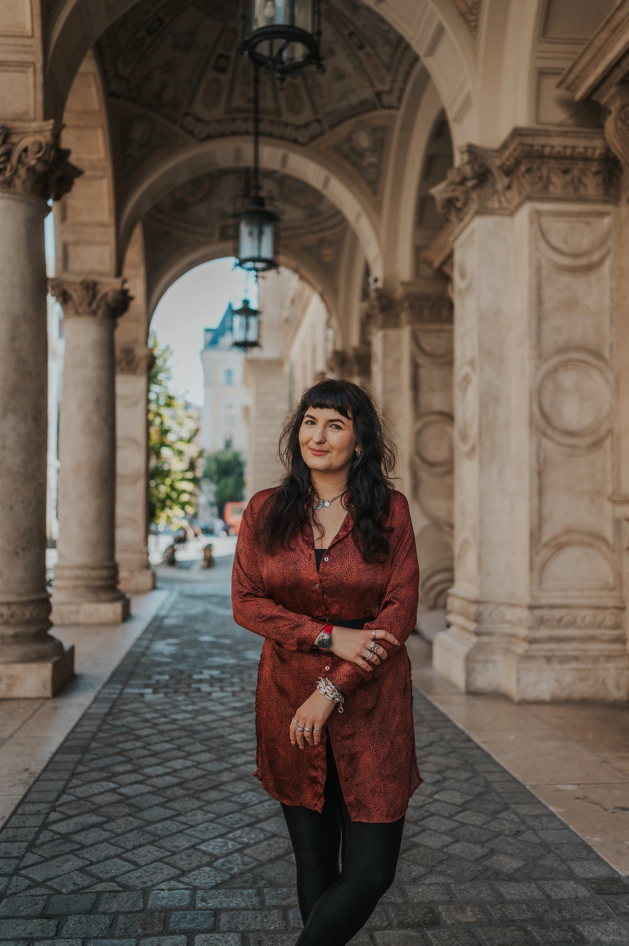 Budapest solo photoshoot: carved arches and a red dress at the Opera The woman standing beneath the Opera’s carved arches with hanging lamps and patterned paving behind her during a solo photoshoot in Budapest.