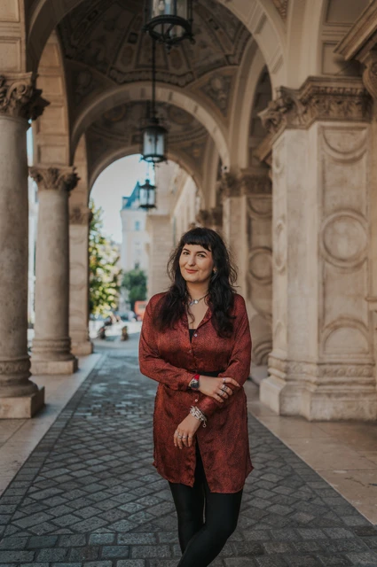 Budapest solo photoshoot: carved arches and a red dress at the Opera The woman standing beneath the Opera’s carved arches with hanging lamps and patterned paving behind her during a solo photoshoot in Budapest.