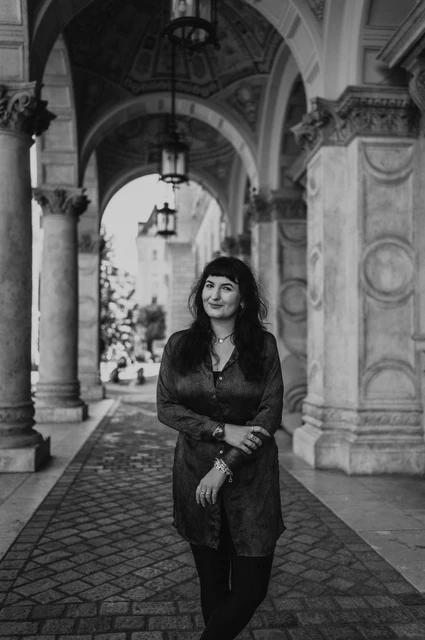 Budapest solo photoshoot: a portrait beneath the Opera’s hanging lamps in black and white Black-and-white photograph of the woman beneath the Opera’s carved stone arcade and hanging lamps during a solo photoshoot in Budapest.