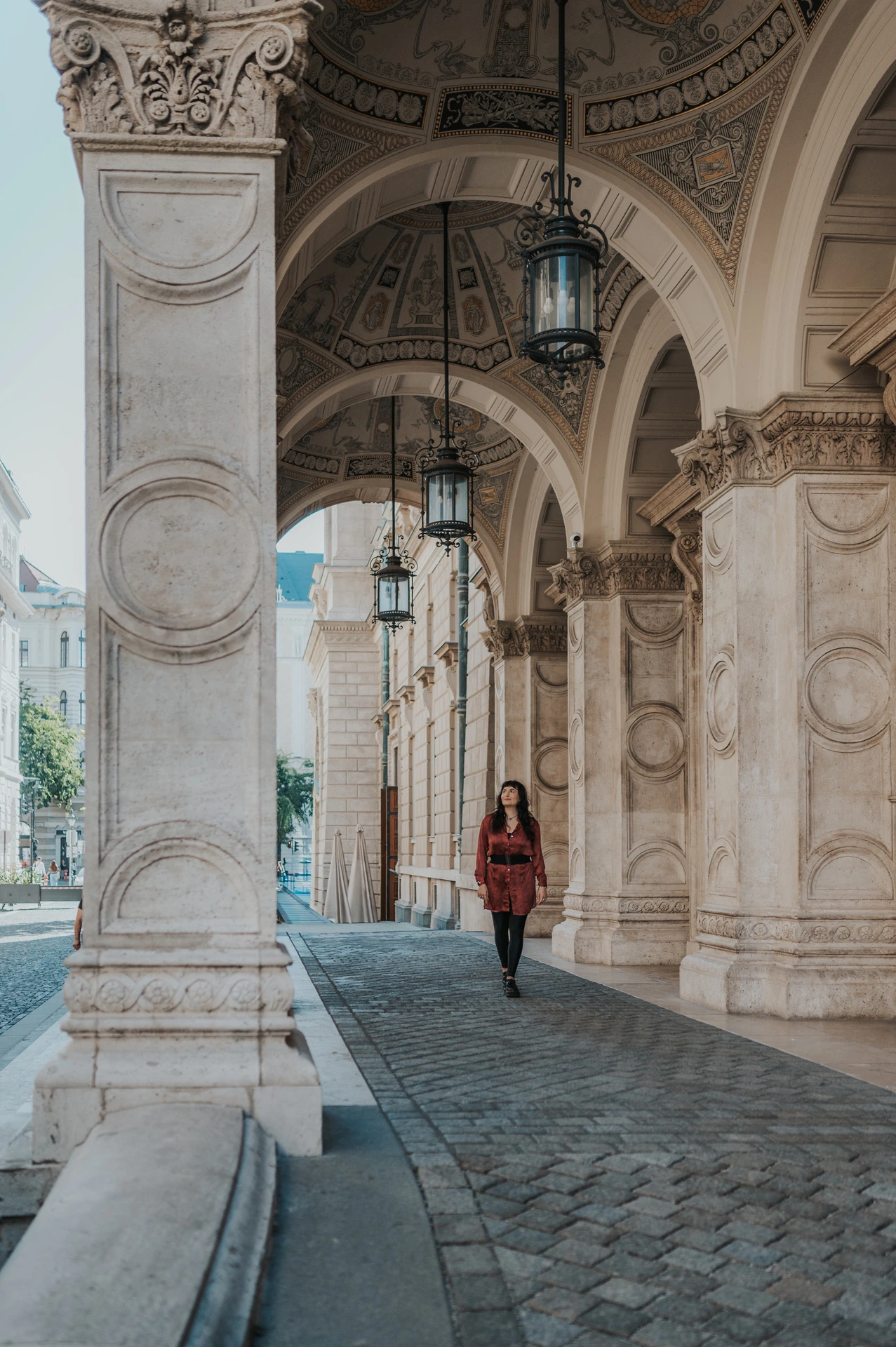 A wide view of the woman beneath the Opera’s ornate stone arcade with its patterned pavement during a solo photoshoot in Budapest.