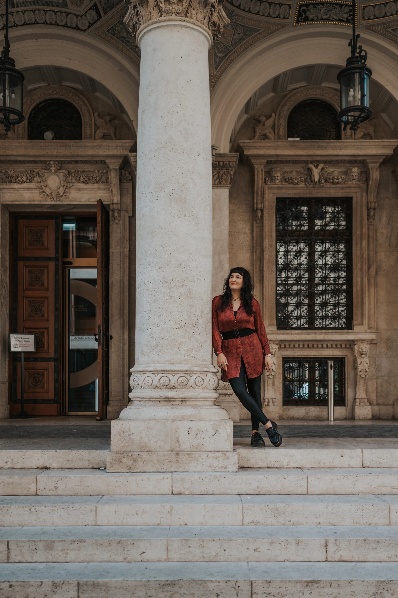 The woman standing beside a tall stone column above the Opera’s broad entrance steps during a solo photoshoot in Budapest.