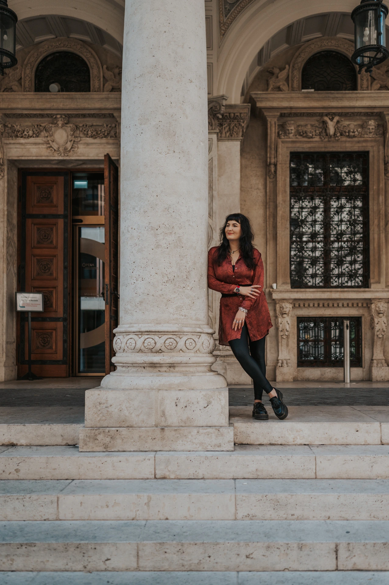 The woman leaning beside a tall column with the Opera’s doors and windows behind her during a solo photoshoot in Budapest.
