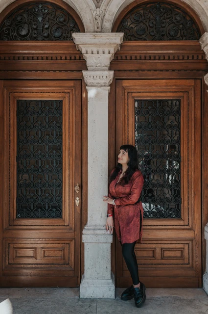 The woman in a red dress standing beside a pale stone column between the Opera’s wooden doors during a solo photoshoot in Budapest.