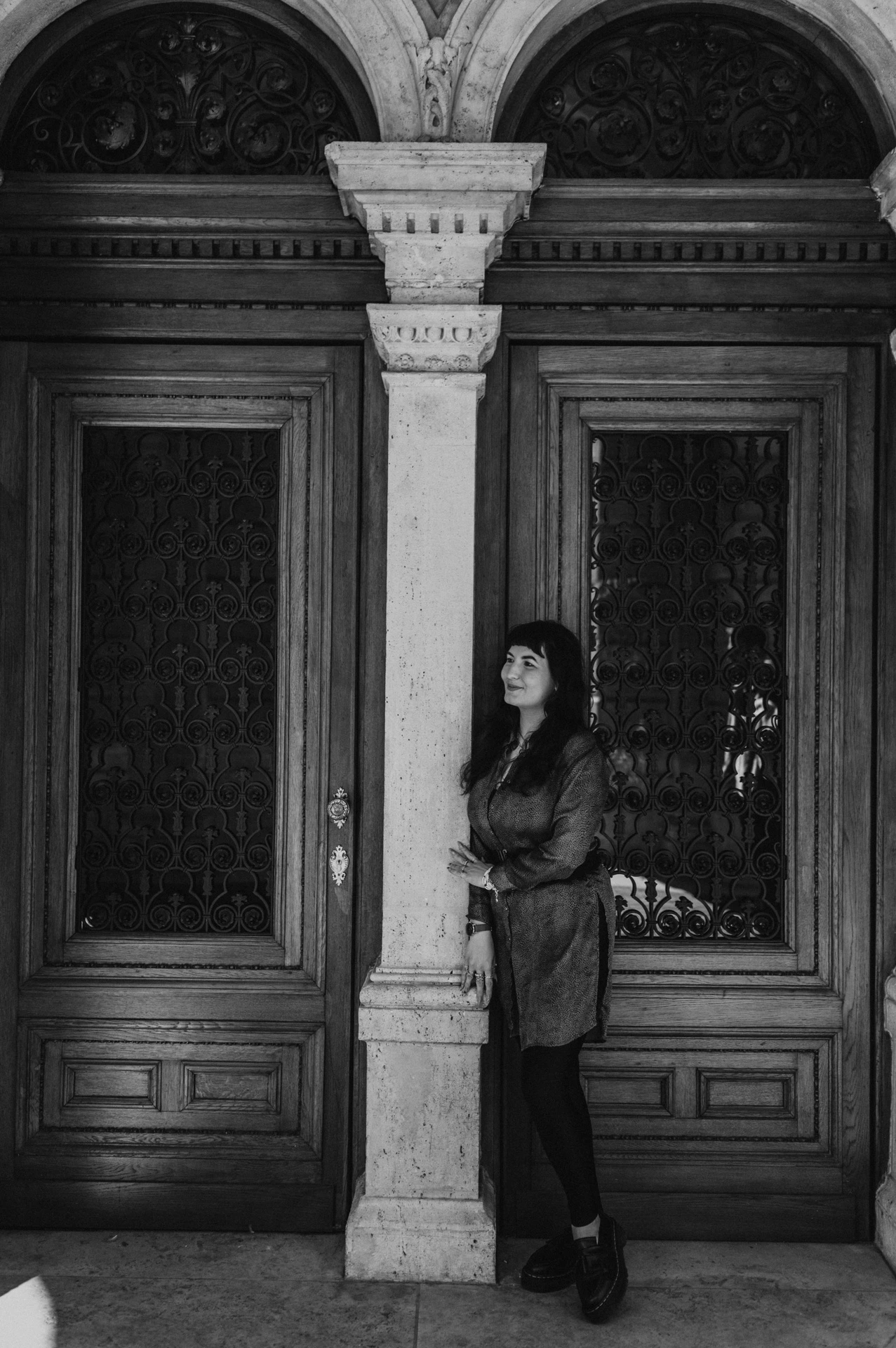 Black-and-white photograph of the woman looking to the side beside the Opera’s carved wooden doors and pale stone column during a solo photoshoot in Budapest.