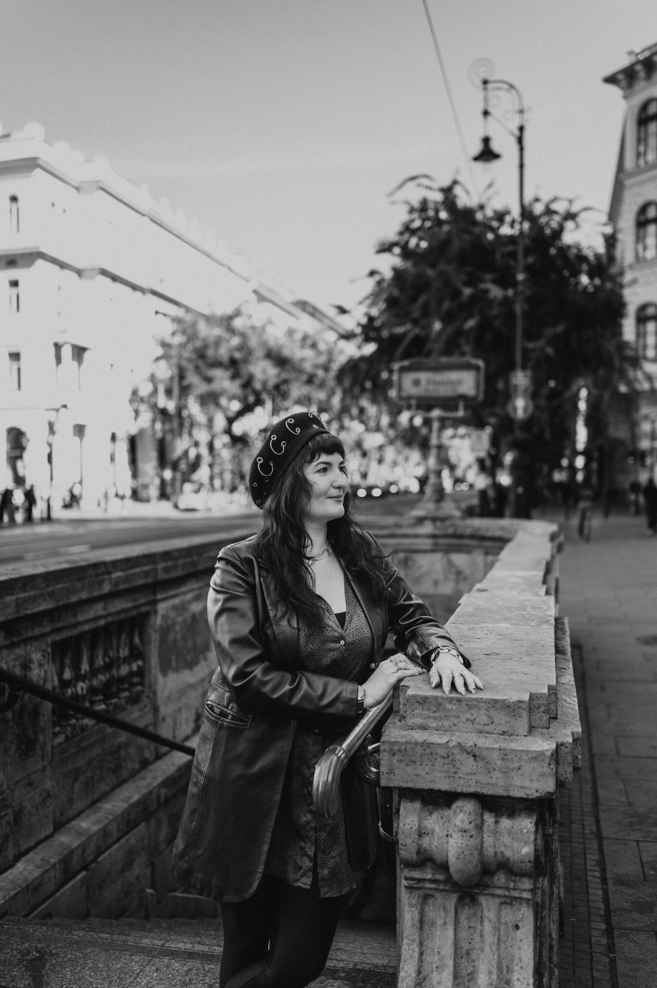 Black-and-white photograph of the woman wearing a dark jacket and beret beside the stone balustrade outside Opera metro station during a solo photoshoot in Budapest.