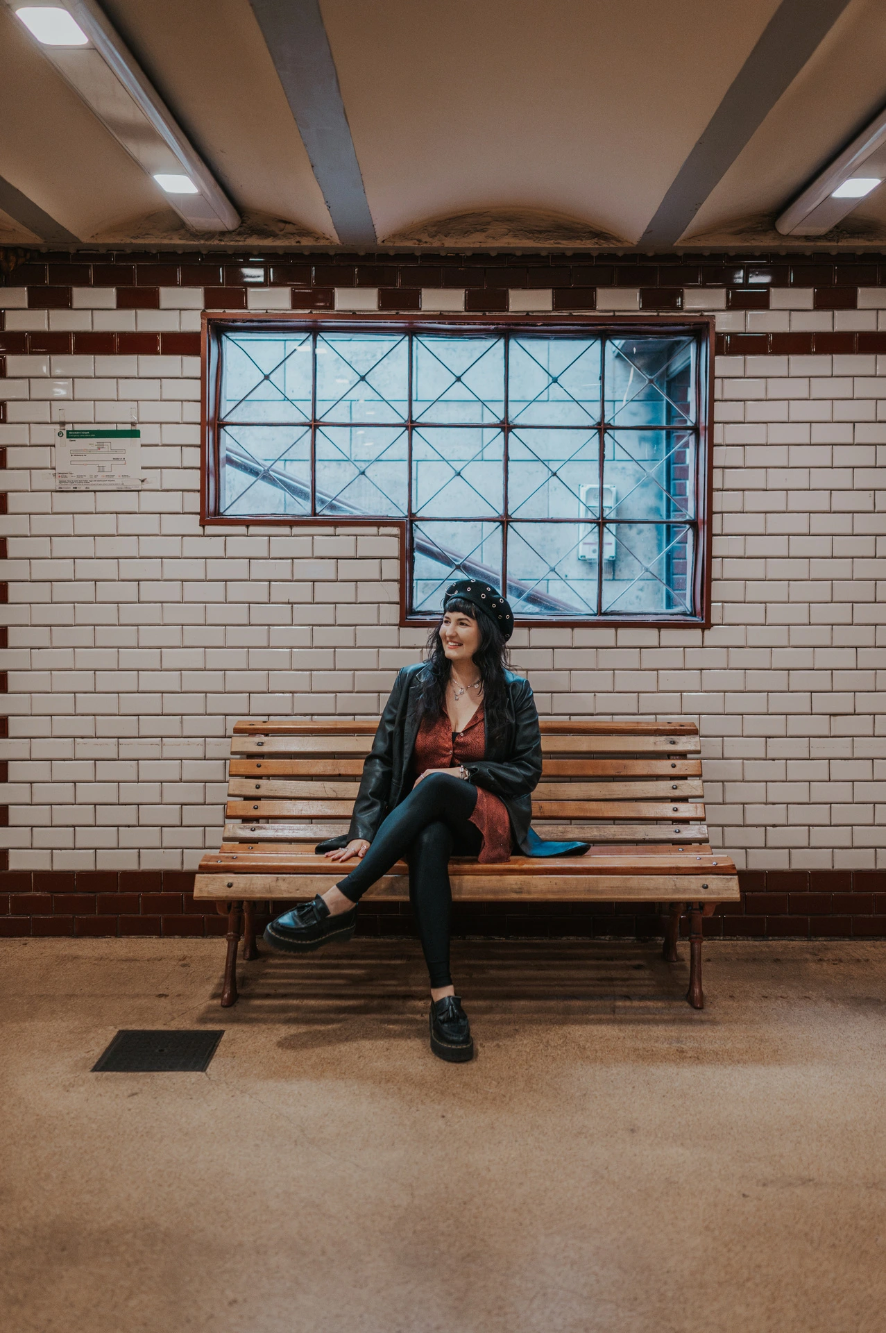The woman sitting on a wooden bench beneath a high window and white tiles at Opera metro station during a solo photoshoot in Budapest.