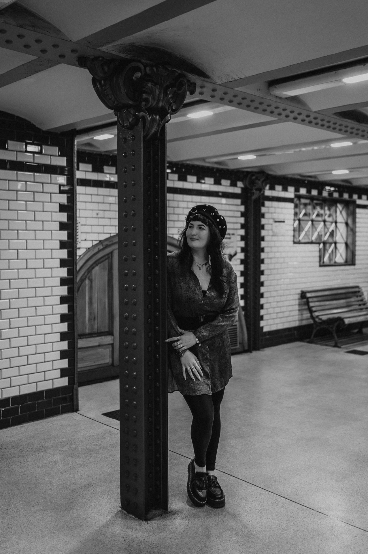 Black-and-white photograph of the woman beside an iron column with white tiles and a wooden bench behind her at Opera station during a solo photoshoot in Budapest.