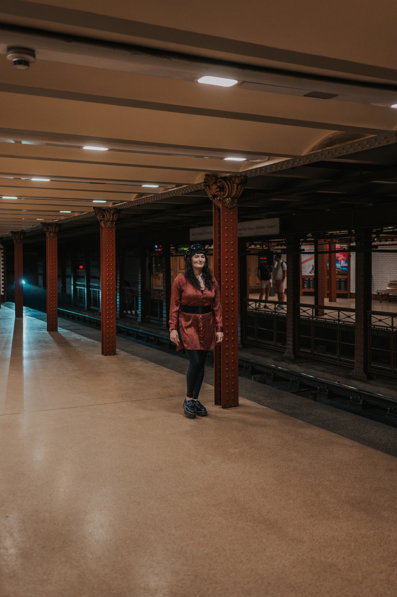 A wide view of the woman on the Opera metro platform with a line of iron columns beside the tracks during a solo photoshoot in Budapest.