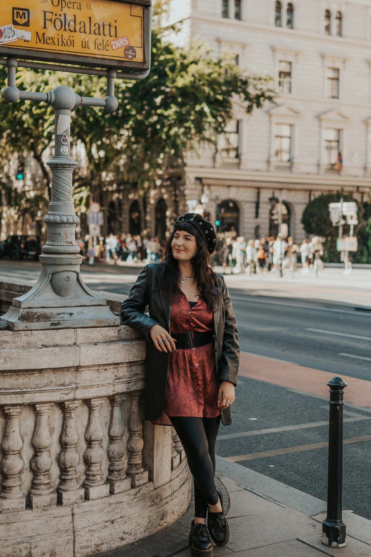 The woman in a red dress, dark jacket and beret beside the yellow Földalatti sign and stone metro balustrade during a solo photoshoot in Budapest.