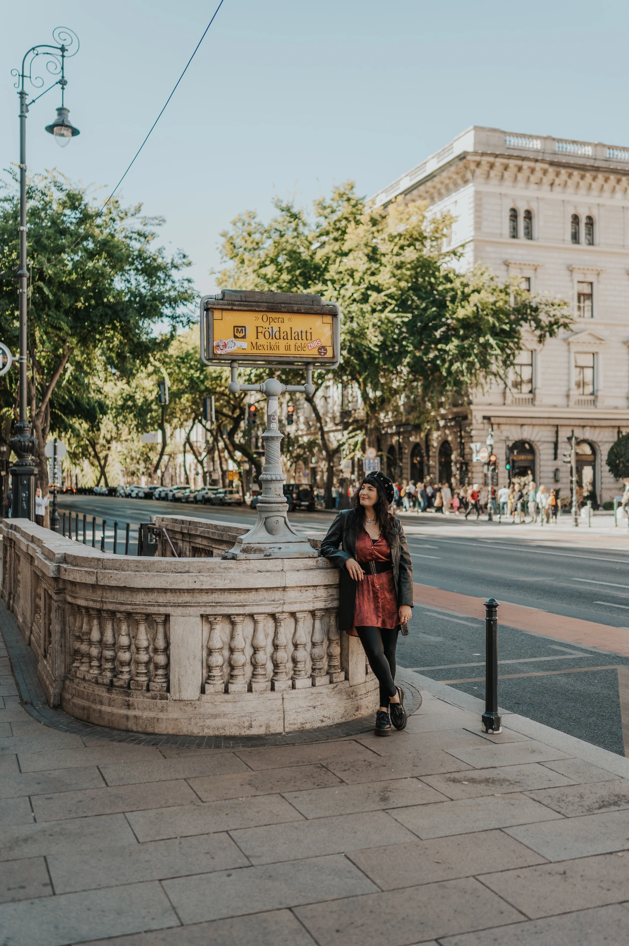 The woman standing beside the curved stone balustrade and yellow Földalatti sign outside Opera station during a solo photoshoot in Budapest.