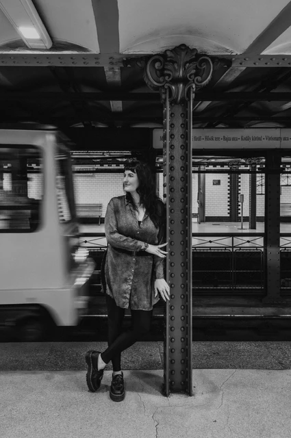 Budapest solo photoshoot: a passing train and an iron platform column in black and white Black-and-white photograph of the woman leaning beside an iron platform column with a moving metro train behind her during a solo photoshoot in Budapest.