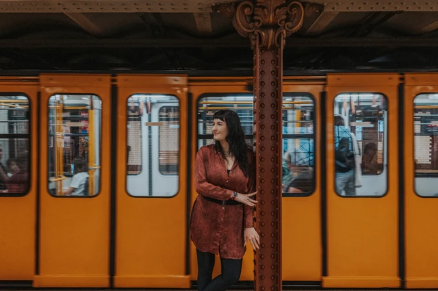 A landscape portrait of the woman beside an iron column with a yellow metro train behind her during a solo photoshoot in Budapest.