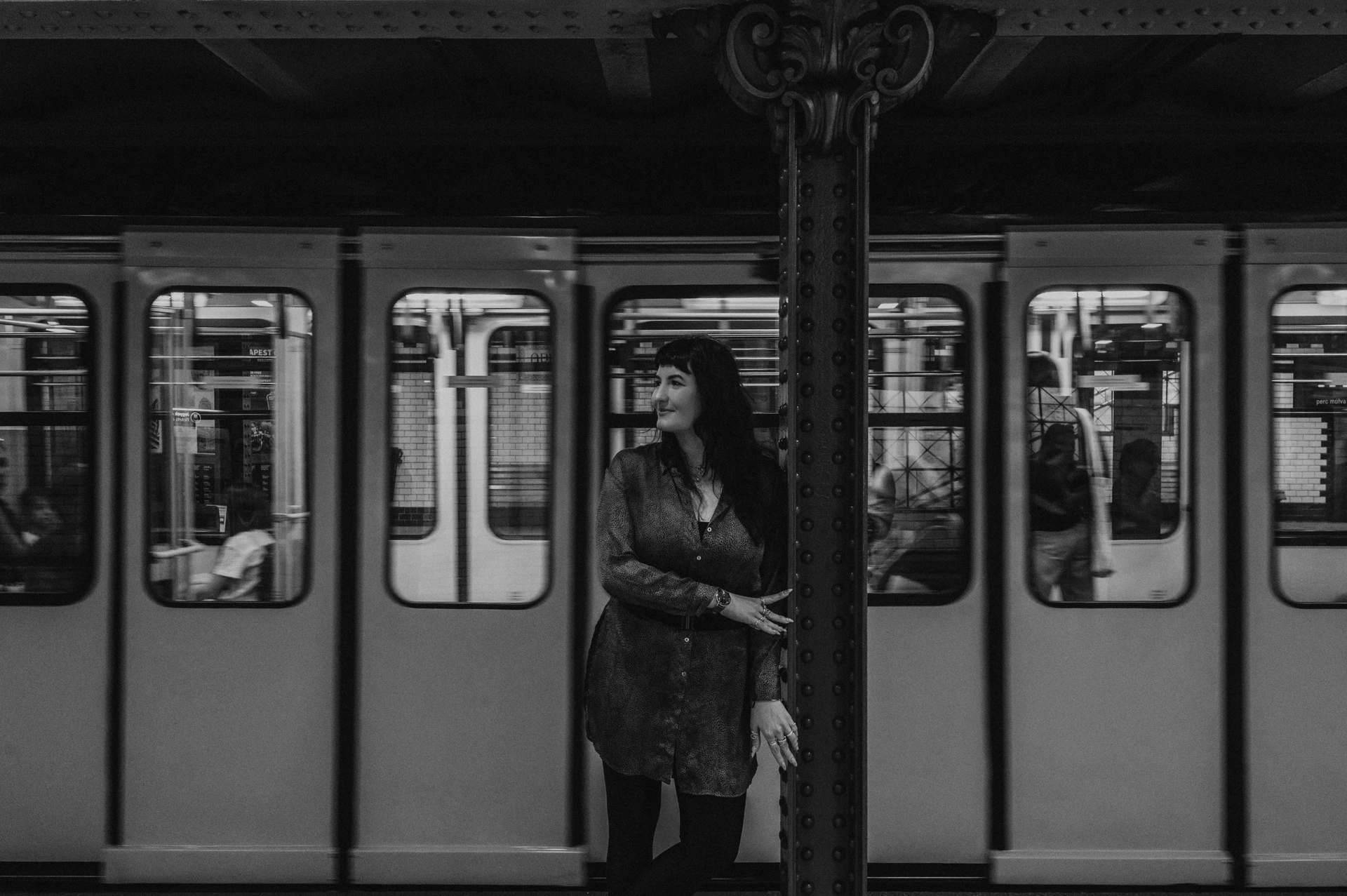 Black-and-white photograph of a landscape portrait of the woman at an iron platform column with a metro train behind her during a solo photoshoot in Budapest.