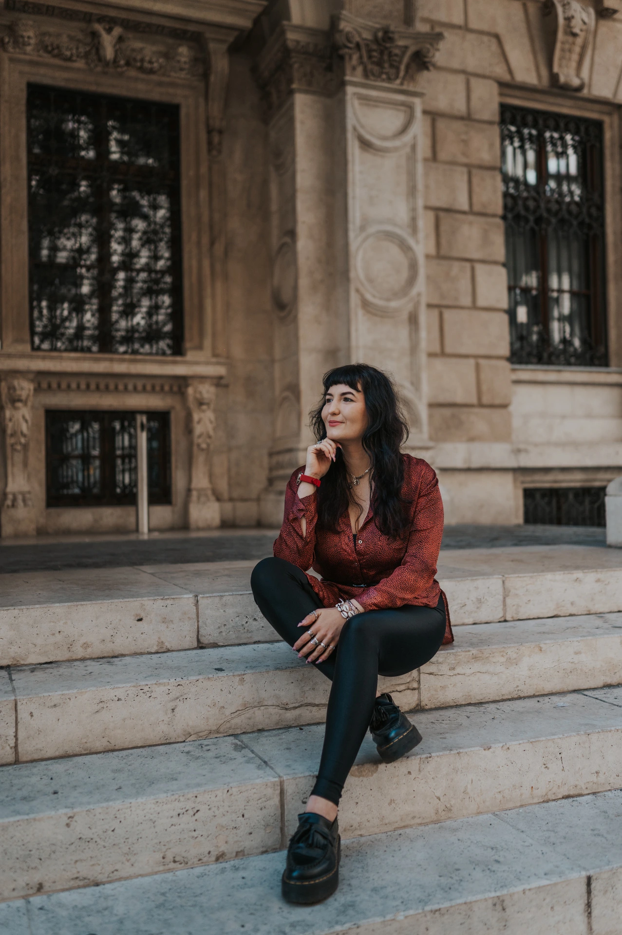 The woman in a red dress sitting on the Opera steps beside the carved stone façade during a solo photoshoot in Budapest.