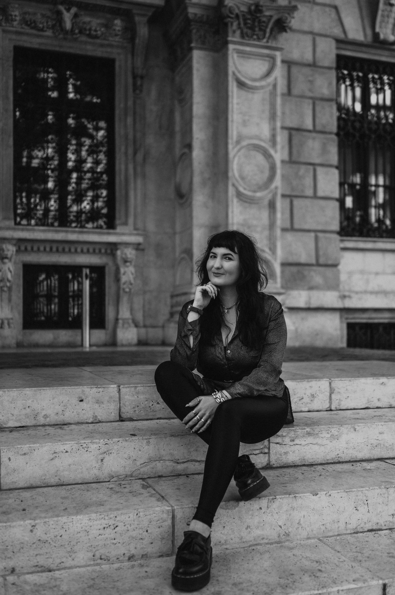 Black-and-white photograph of the woman sitting on the Opera steps with carved stonework and a tall window behind her during a solo photoshoot in Budapest.