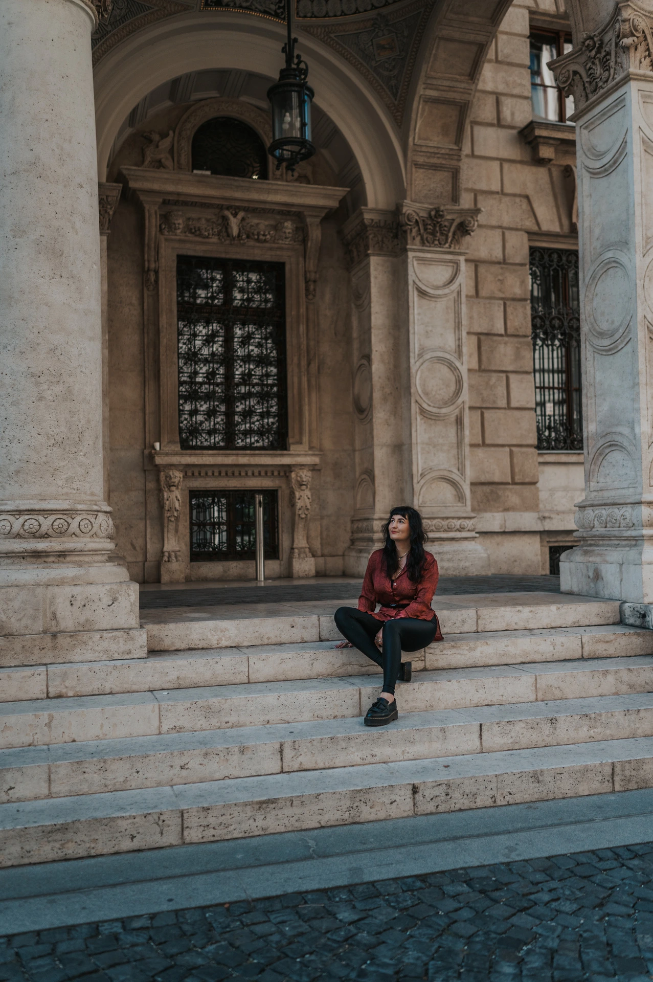 The woman sitting on the broad stone steps beneath an arched Opera window during a solo photoshoot in Budapest.