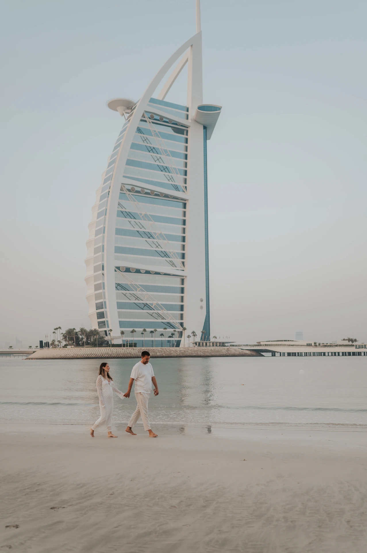 An expecting couple walks across wet sand in Dubai with Burj Al Arab behind them during a maternity photoshoot at golden hour.