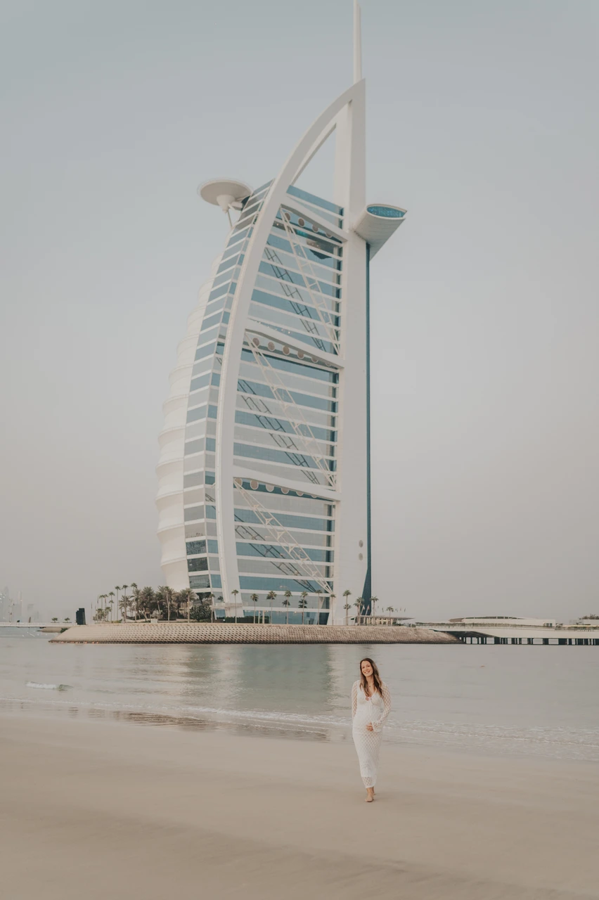 Burj Al Arab maternity photoshoot Dubai - hero portrait A pregnant woman stands on Dubai beach with Burj Al Arab visible behind her during a maternity photoshoot at soft golden hour.