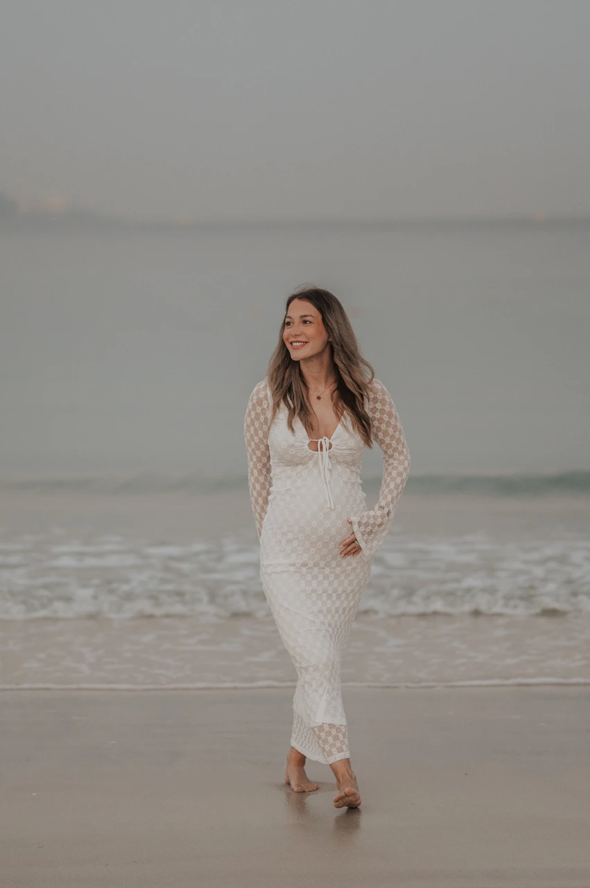 Burj Al Arab maternity photoshoot Dubai - solo white dress A solo maternity portrait shows an expecting mother who stands in a white dress on Dubai beach with Burj Al Arab blurred behind during a maternity photoshoot.