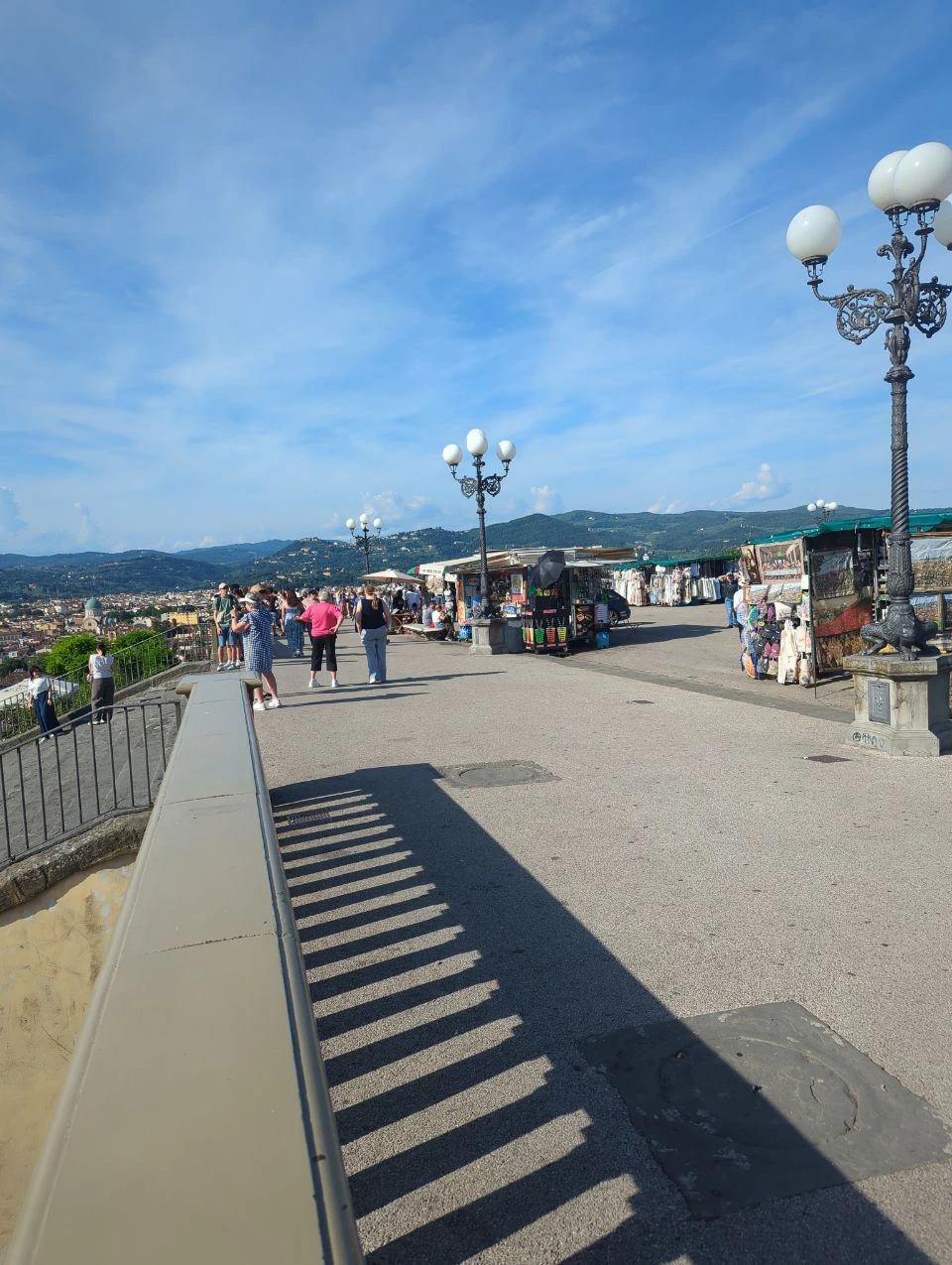 Busy Florence Hilltop Viewpoint Visitors gather near stalls and ornate lamps at a busy Florence hilltop viewpoint, with the city and hills beyond.