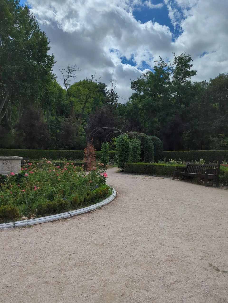 Family Photoshoot in Madrid With Kids - Planned Garden Break A bench sits beside a curved gravel path and rose beds in the Campo del Moro gardens in Madrid.