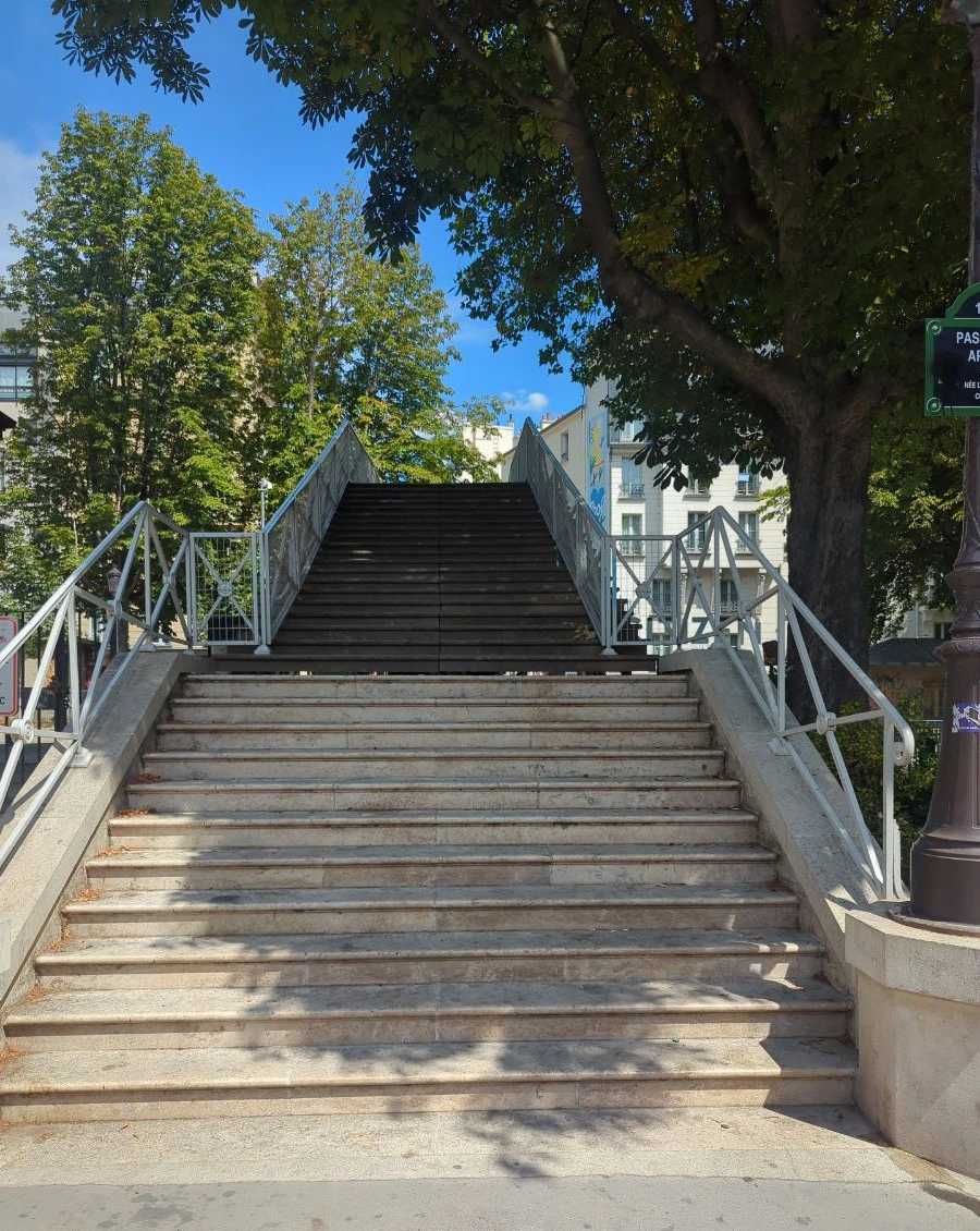 Paris Photoshoot - Canal Saint-Martin Footbridge A broad metal footbridge rises beneath mature trees beside Canal Saint-Martin, adding urban structure and a connected walking route to a Paris photoshoot.