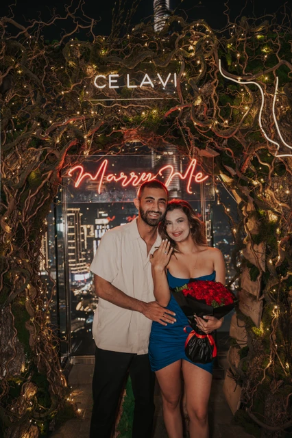 A newly engaged couple smiles at CE LA VI in Dubai, with the engagement ring visible and a bouquet of red roses in hand.