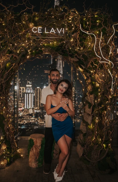 A couple poses under the CE LA VI sign on a Dubai rooftop after their nighttime proposal, with city lights behind them.