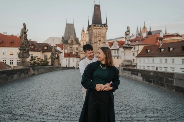A couple hold each other on Charles Bridge, with the Lesser Town Bridge Towers and red rooftops behind them.