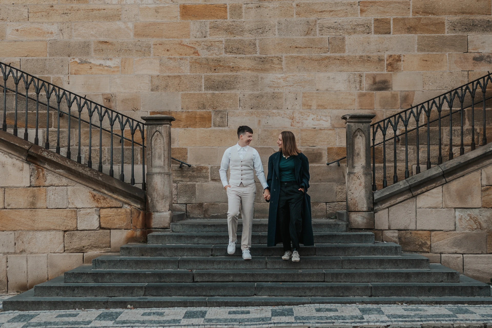 A couple hold hands on the Kampa staircase, between matching iron railings and the warm stone walls of Charles Bridge.