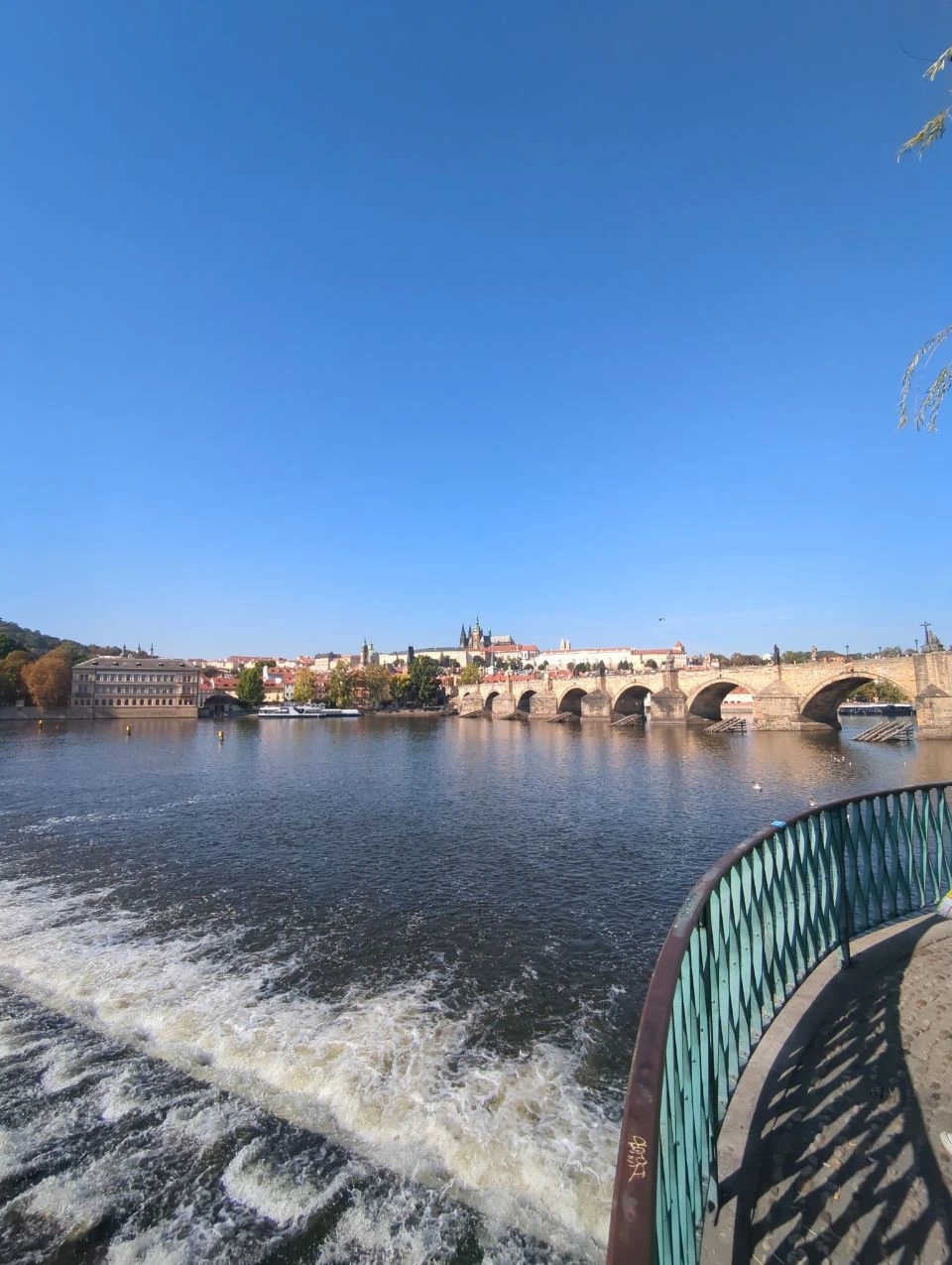 Charles Bridge Viewpoints - Riverside Weir and Prague Castle Water flows over a weir beside a curved railing, with Charles Bridge and Prague Castle across the Vltava.