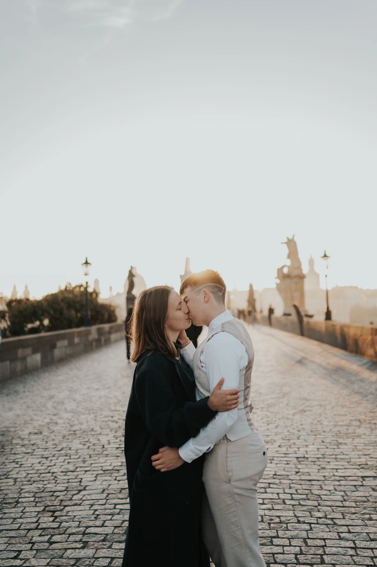 Charles Bridge Photos - Couple in Morning Sunlight A couple kiss on Charles Bridge as low morning sunlight shines behind them, with statues and lamps along the walkway.