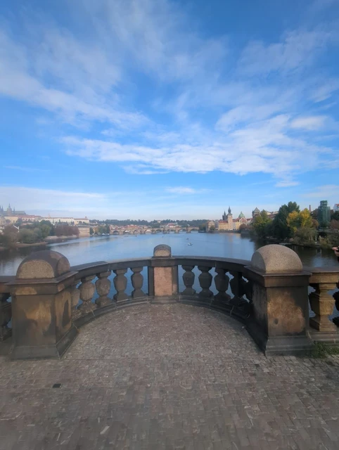 A curved stone balustrade on Legion Bridge overlooks the Vltava, with Charles Bridge and Prague’s towers in the distance.