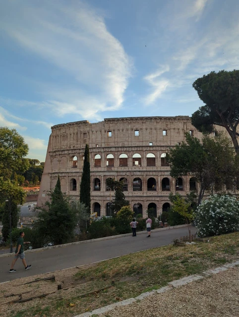 Colosseum from an Elevated Garden Viewpoint The Colosseum rises beyond a sloping path, trees and flowering shrubs under a softly clouded blue sky in Rome.