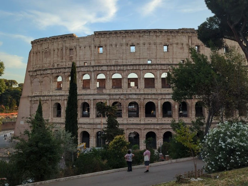 Two visitors stand on a landscaped path below the Colosseum, framed by tall trees and white flowers in Rome.