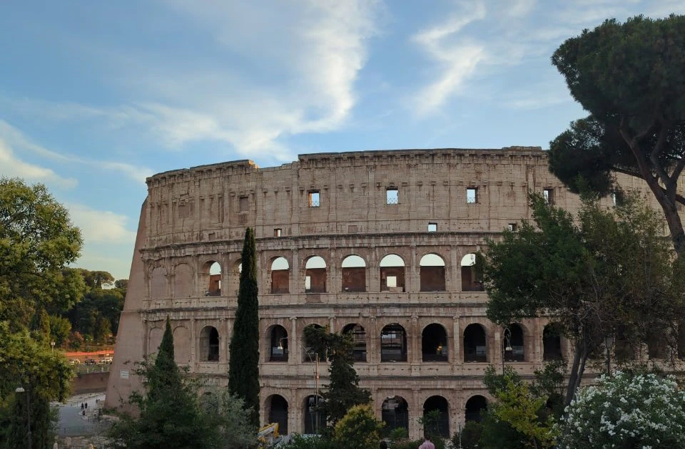 A quiet elevated Rome photoshoot viewpoint frames the Colosseum between trees beneath a softly clouded blue sky.