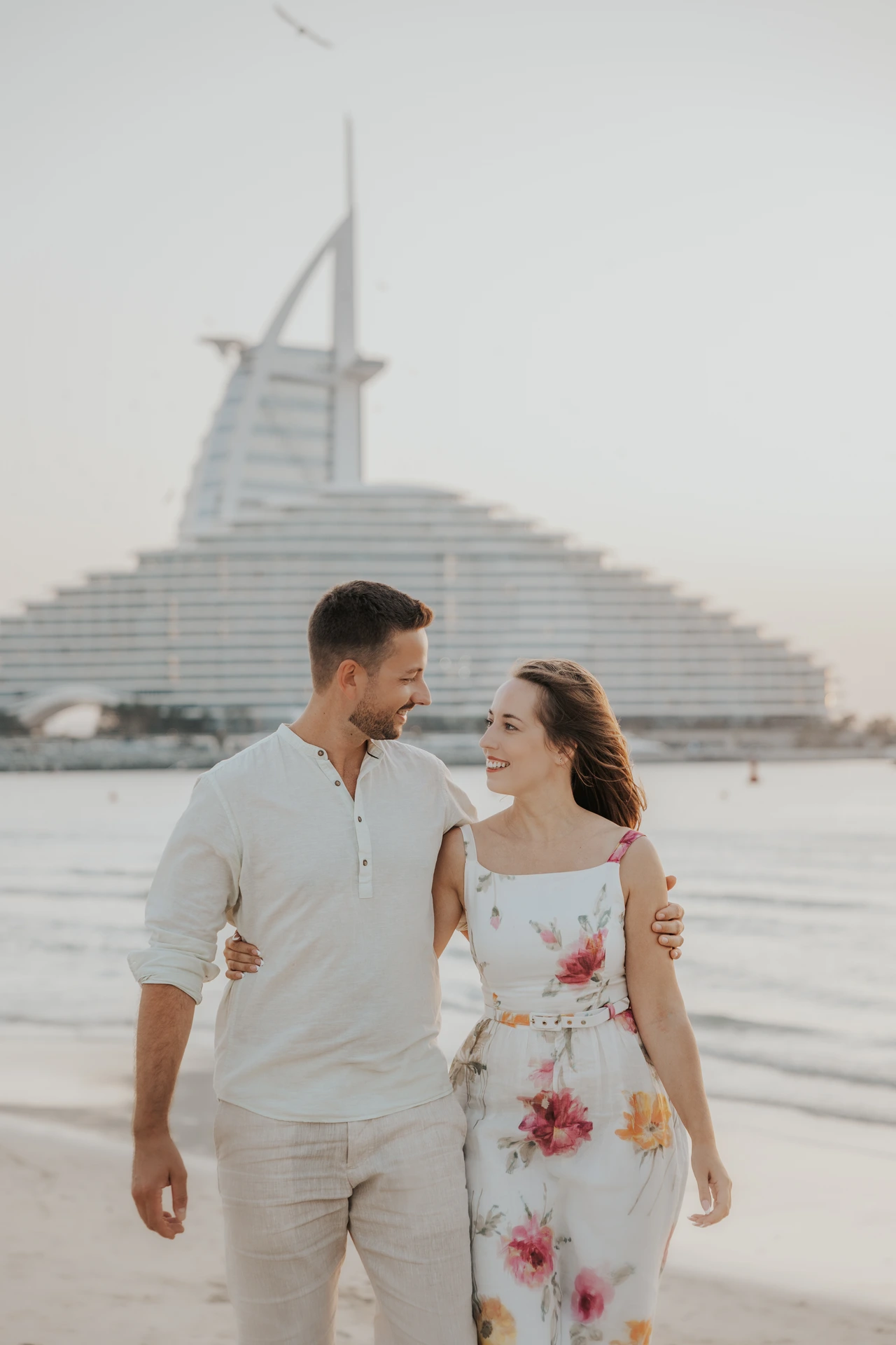A smiling couple stands close together on a Dubai beach with Burj Al Arab in the background.