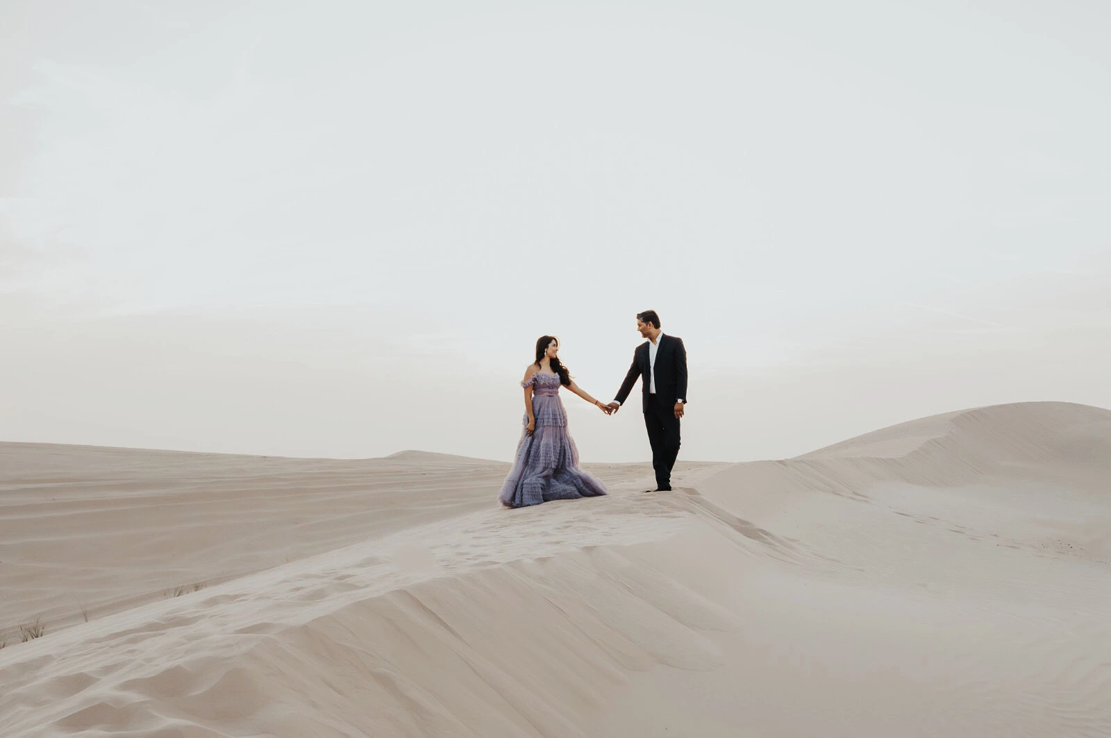 A wide shot of a couple walking hand in hand across Dubai desert dunes at sunset.