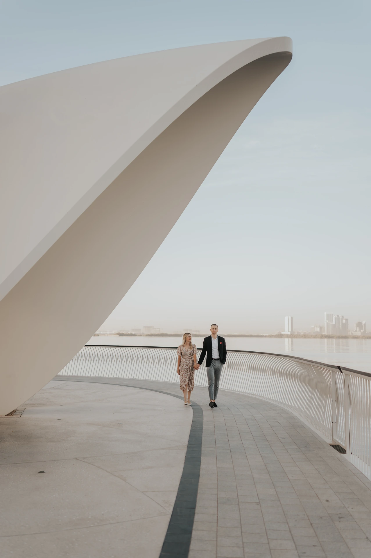 A couple walks along a modern waterfront promenade in Dubai with clean architectural lines and skyline in the distance.