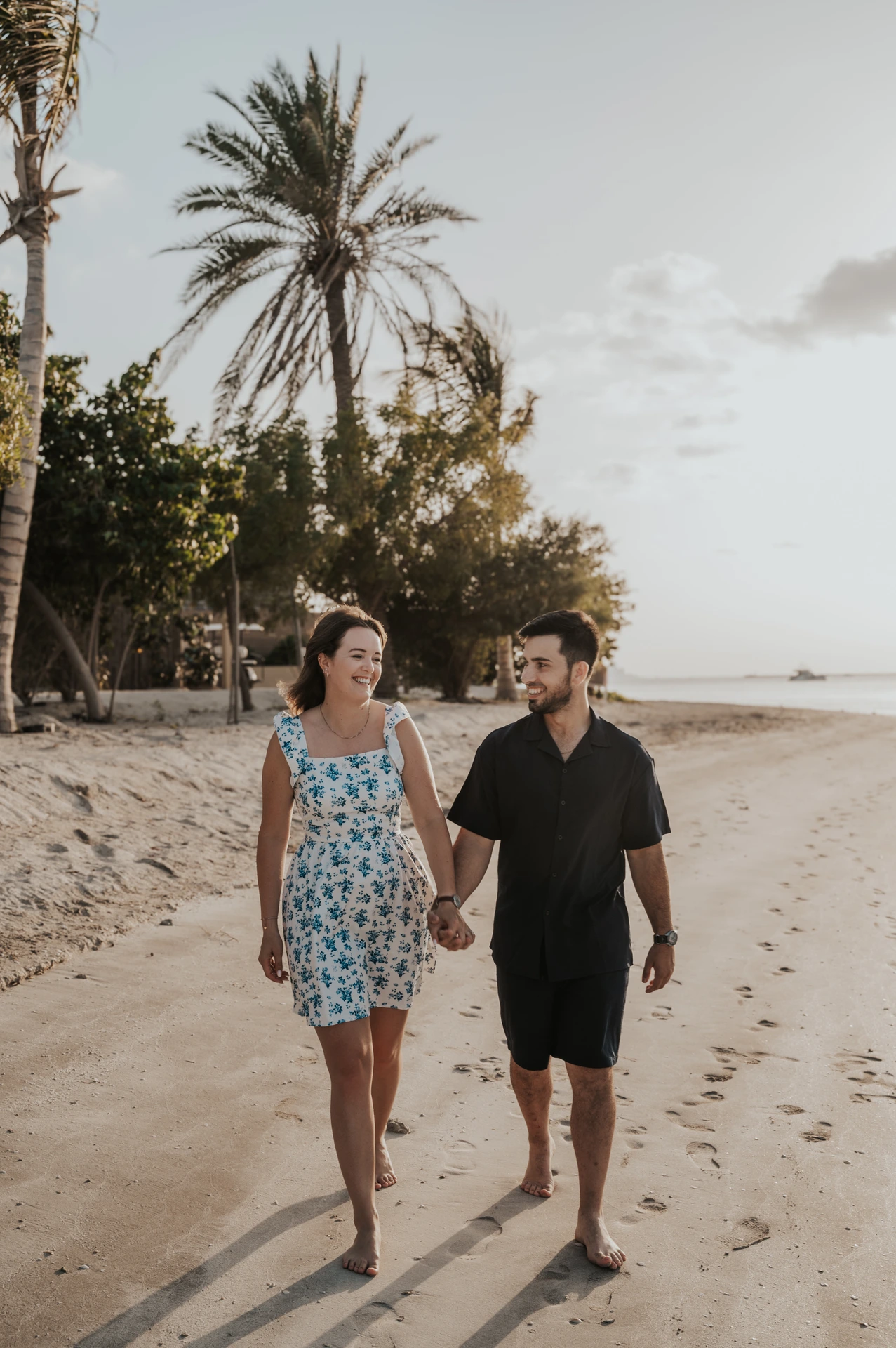 A couple walks hand in hand along a quiet Dubai beach in soft early light during a relaxed photoshoot.