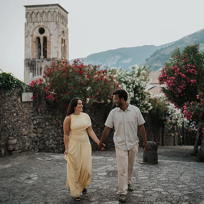 Couple walking beside a flowering stone wall with a bell tower behind them