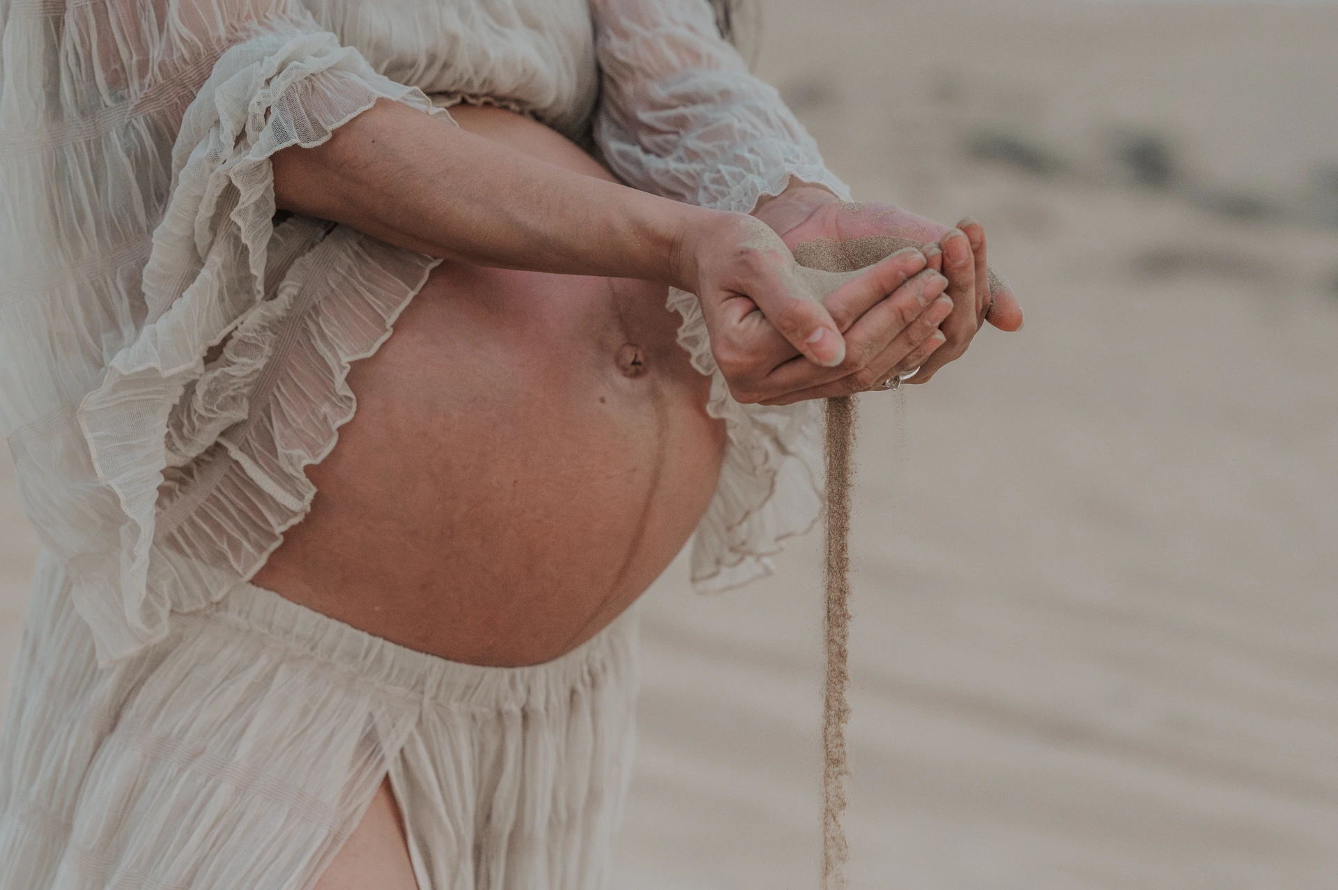 Close-up of a pregnant belly and hands pouring sand in Dubai desert during a serene maternity photoshoot.