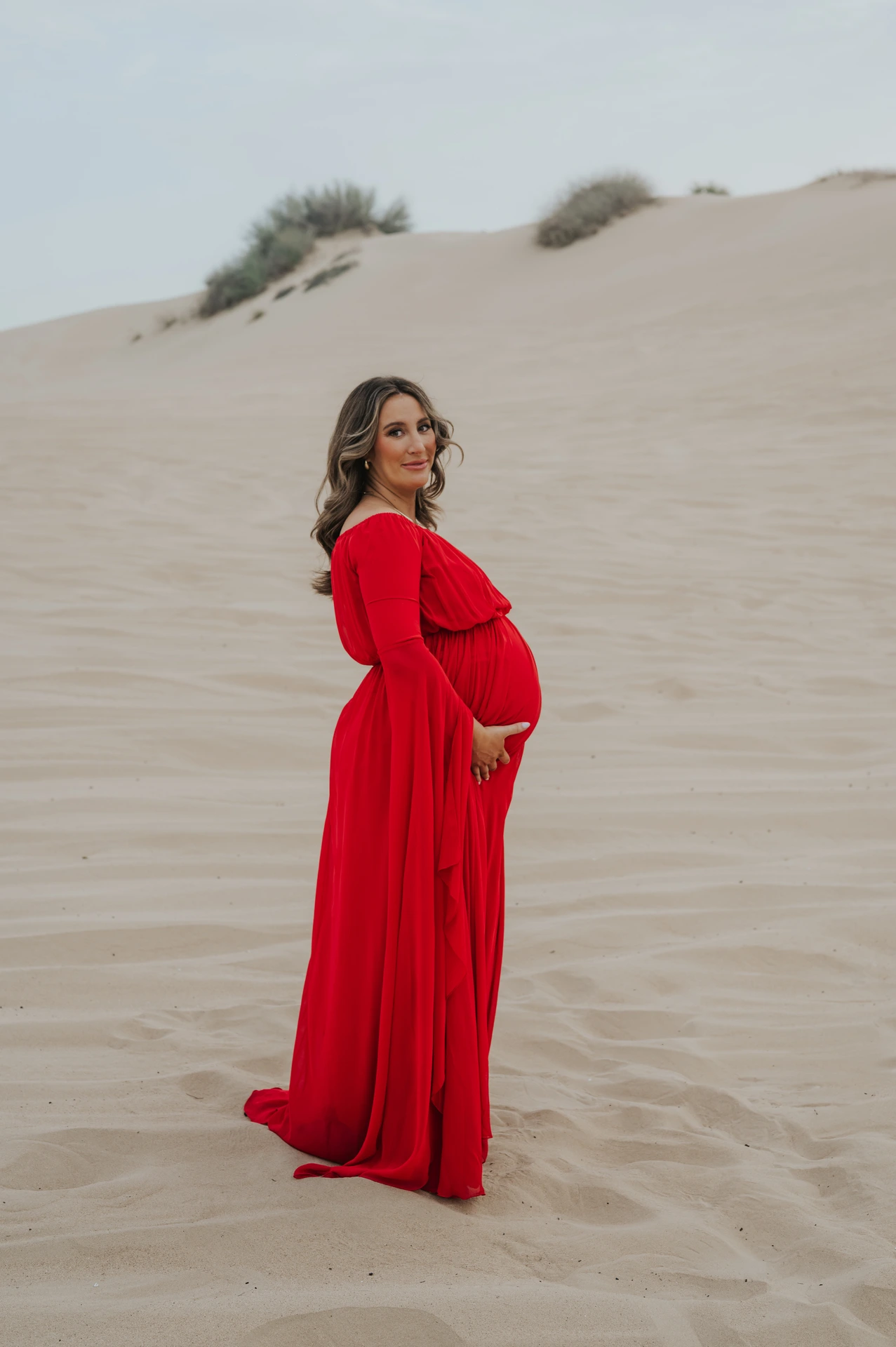 Pregnant woman in a flowing red dress stands on Dubai desert dunes during a serene maternity photoshoot session.