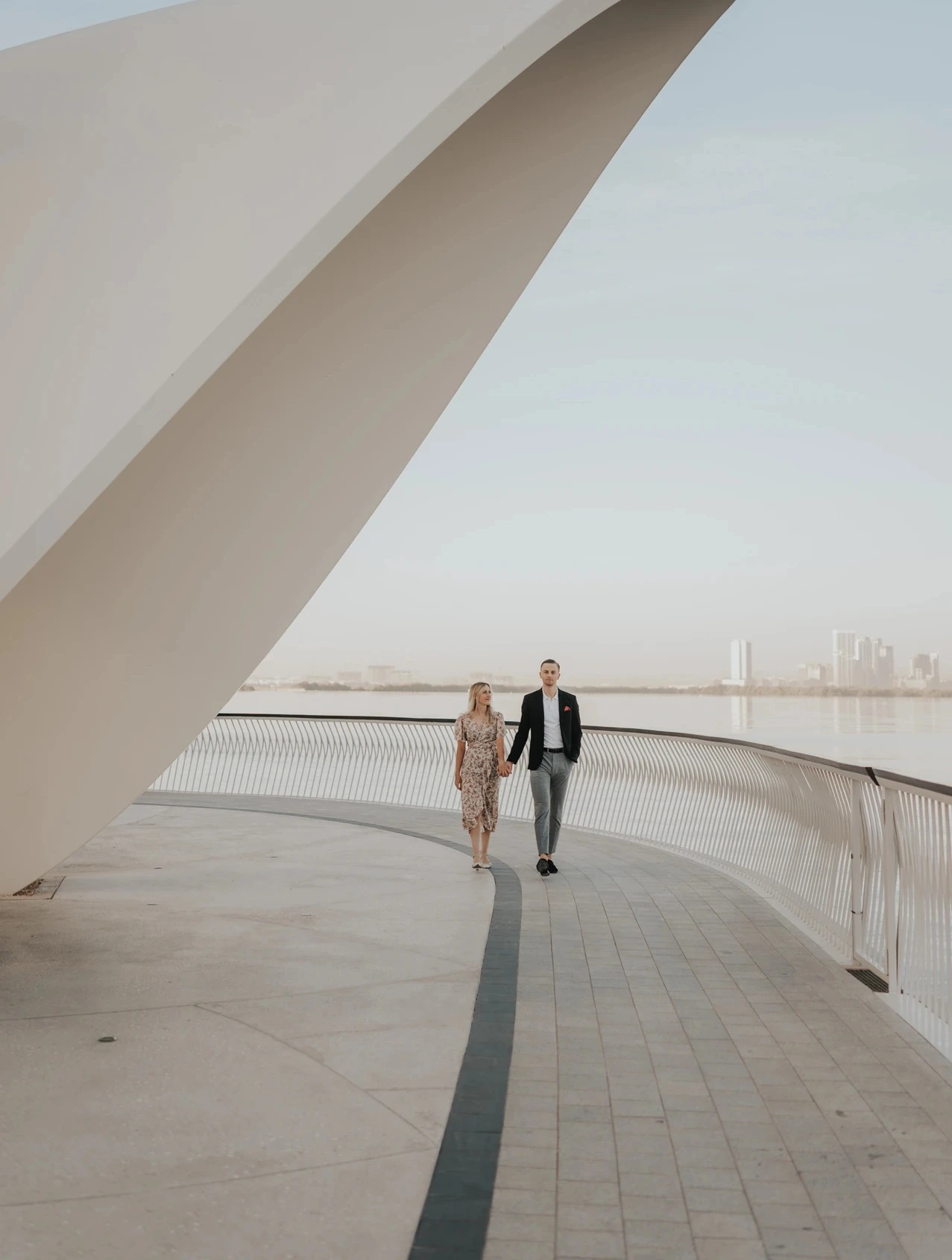 A couple walks beneath a modern architectural frame near the Dubai waterfront with skyline buildings in the distance.