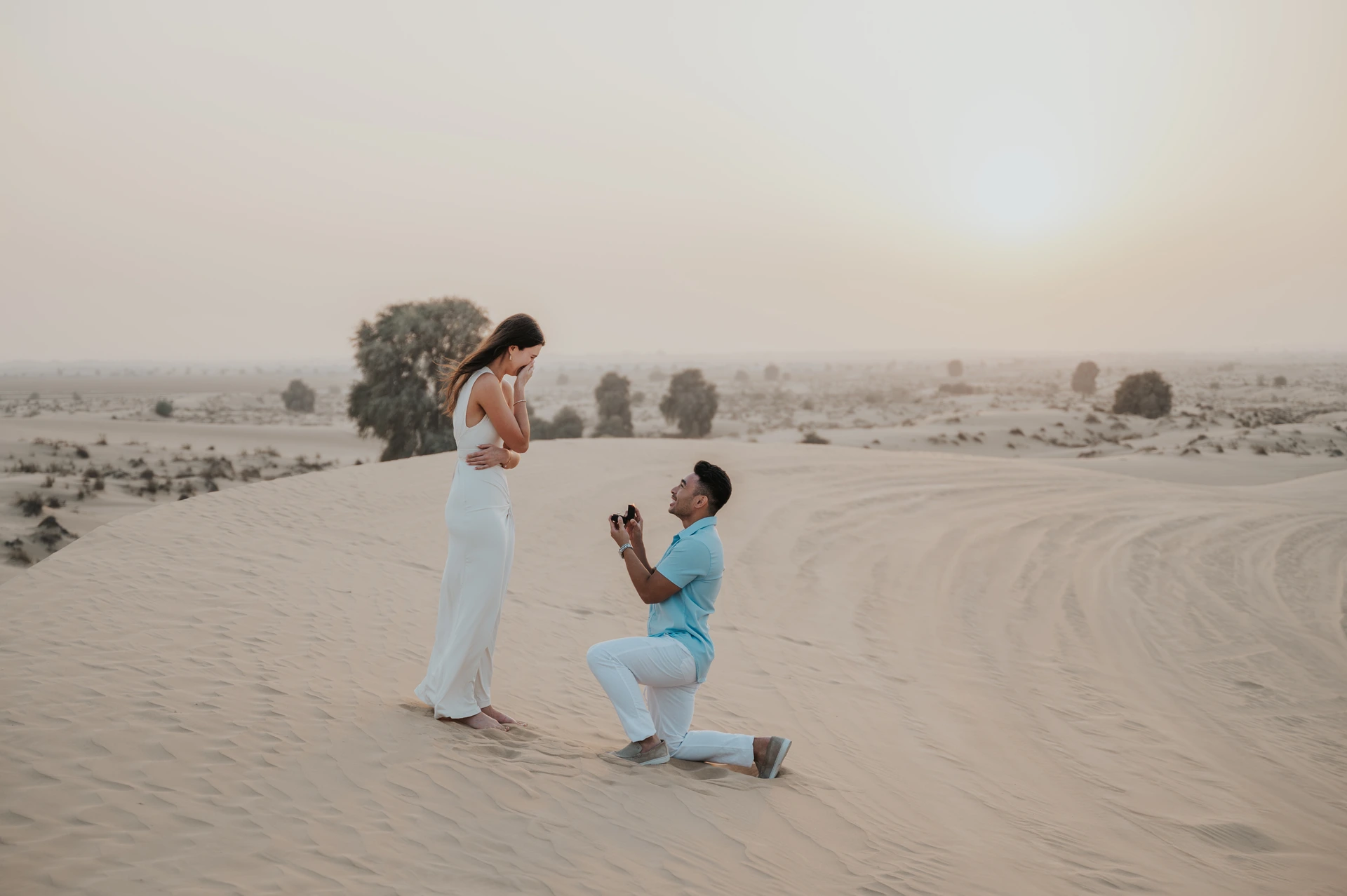 A man kneels to propose on open Dubai dunes while his partner smiles in soft sunset light.