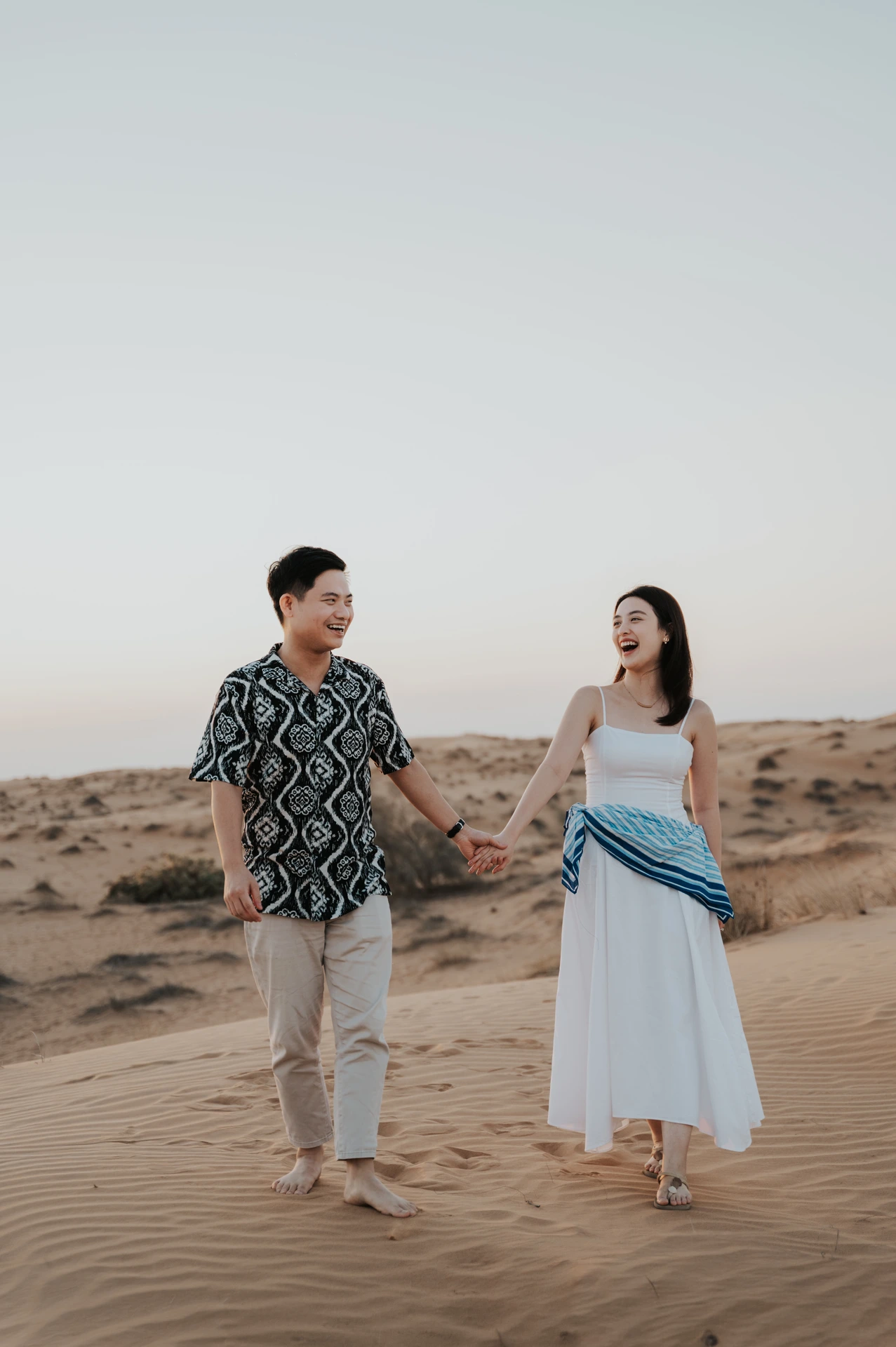 A couple laughs while walking barefoot on Dubai dunes during golden-hour proposal portraits.