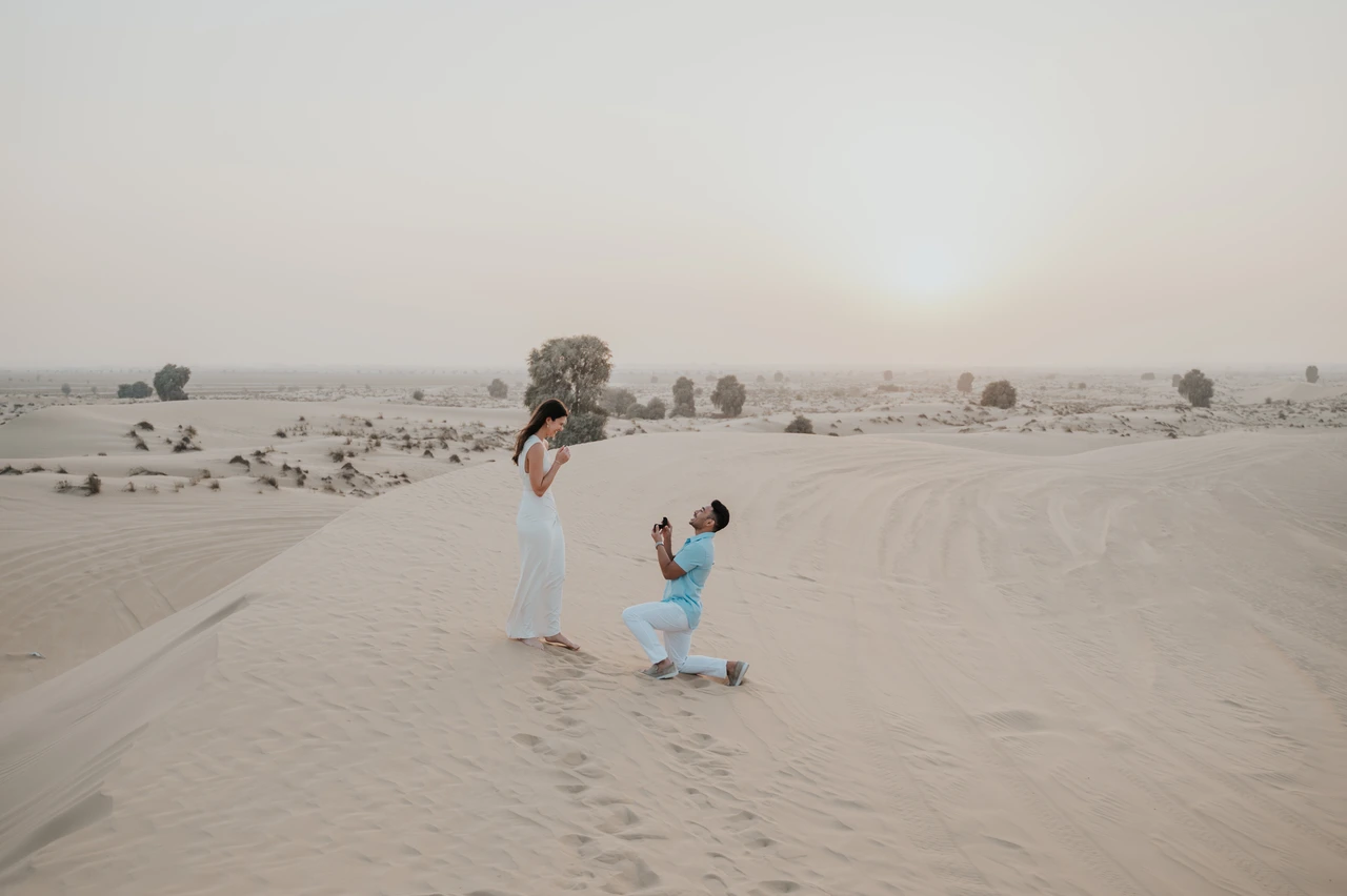 Dubai Proposal - Desert Bouquet Question A man kneels with flowers on pale desert sand for a Dubai proposal as his partner stands nearby.